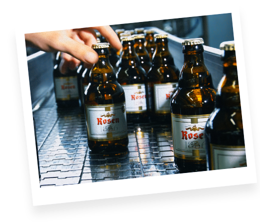 mehrere Flaschen 0,33 l Rosen Pils auf einem Fließband. Ein Mitarbeiter greift eine Flasche mit der linken Hand.